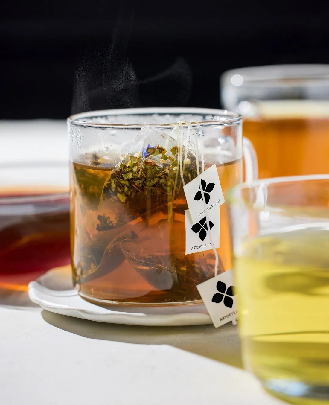 A steaming glass of Art of Tea with sachets steeping, teacups alongside